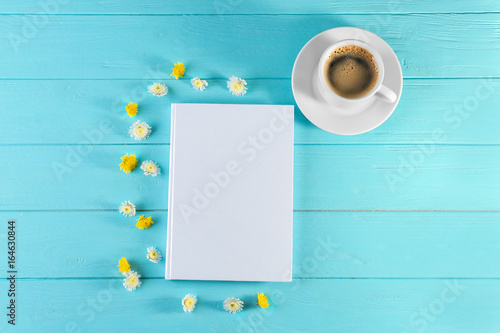 Book with blank white cover, cup of coffee and beautiful flowers on wooden background