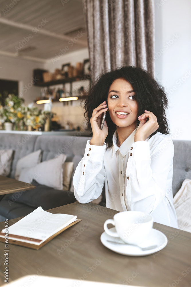 Young African American girl sitting in restaurant and talking on her mobile phone. Lady with dark curly hair sitting in cafe with cup of coffee and menu on table. Smiling lady in white shirt Young African American girl sitting in restaurant and talking on her mobile phone. Lady with dark curly hair sitting in cafe with cup of coffee and menu on table. Smiling lady in white shirt