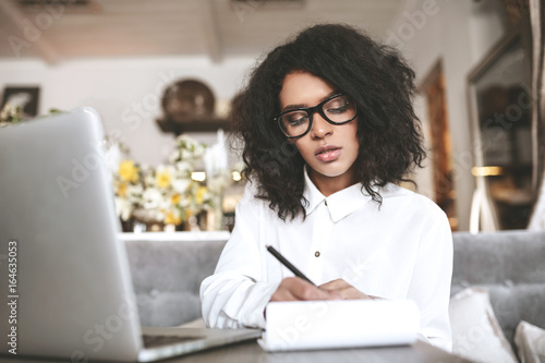 Young girl in glasses sitting in restaurant and thoughtfully writing in her notebook. Pretty African American girl working at cafe with laptop. Portrait of lady with dark curly hair making notes