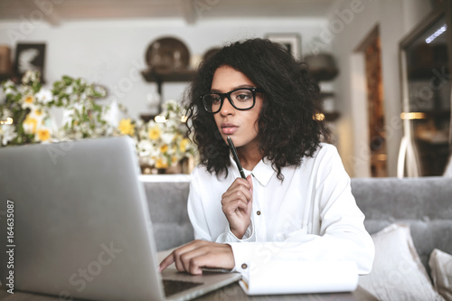 Young African American girl working on laptop in restaurant. Pretty girl with dark curly hair sitting in cafe with laptop. Portrait of lady in glasses thoughtfully looking in laptop with pen in hand