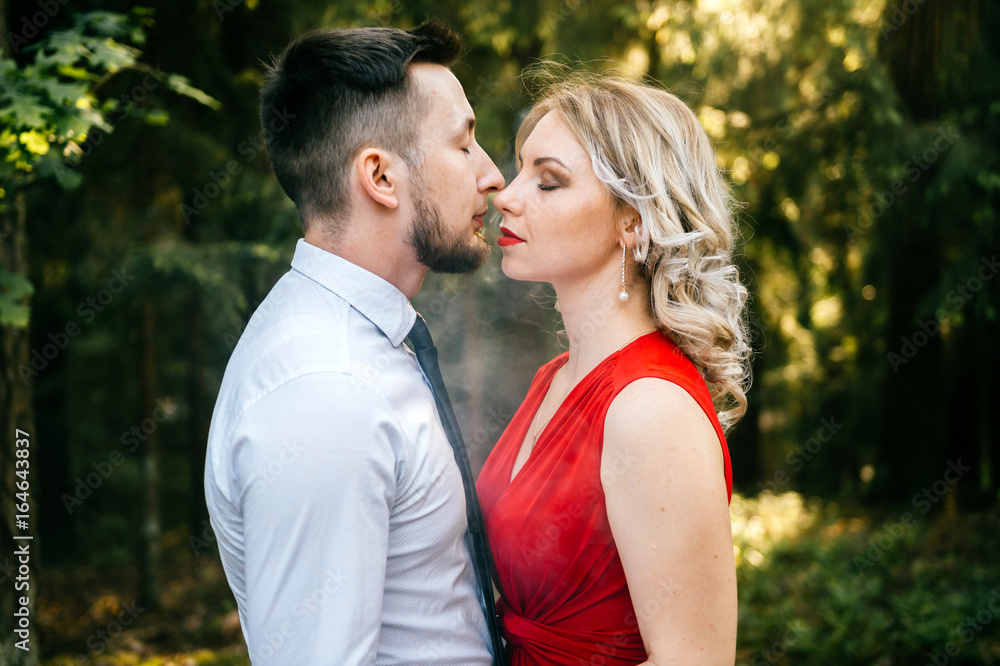 Fototapeta premium Closeup of couple of lovers kissing with smoke outdoor. Businessman and young blonde girl with red lips, curly hair and red dress with decolette. Smokers in forest in summer sunny day. Sensual moment