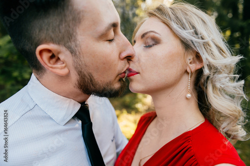 Closeup of couple of lovers kissing with smoke outdoor.  Businessman and young blonde girl with red lips, curly hair and red dress with decolette. Smokers in forest in summer sunny day. Sensual moment © benevolente
