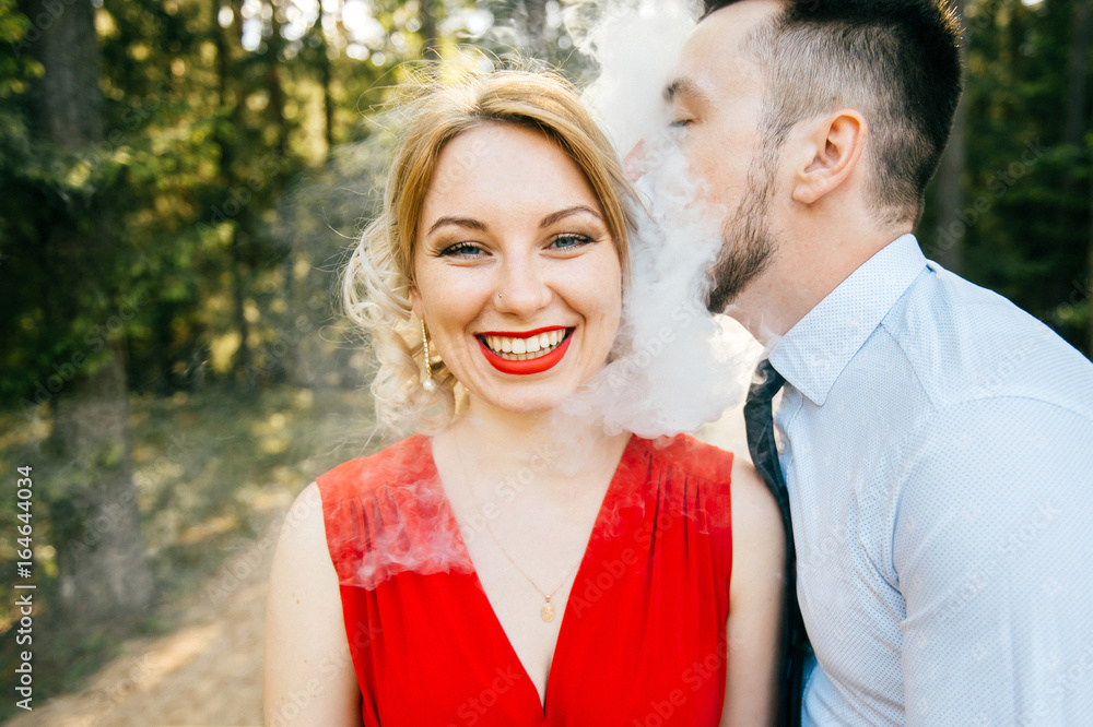 Fotka „Smoking man in business clothes kissing young blonde girl with red  lips, dress with decolette. Clouds of thick smoke. Couple of lovers  outdoor. Smiling and laughing woman. E-cigarette.“ ze služby Stock