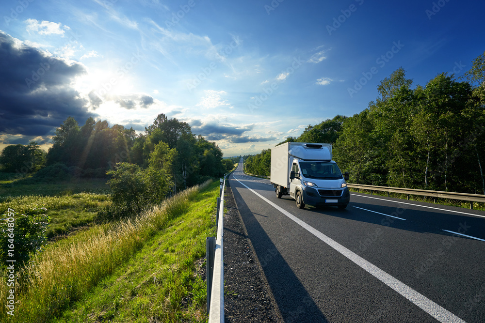 Delivery car driving on the asphalt road between a forest in a rural landscape at sunset