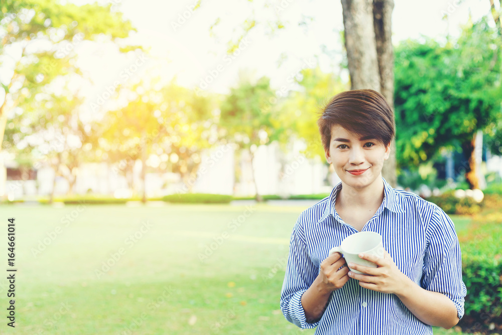 Beautiful short hair woman standing in the green yard and drinking a favorite beverage in white cup for resting between her outdoor working business in the park. Beautiful short hair woman standing in the green yard and drinking a favorite beverage in white cup for resting between her outdoor working business in the park.