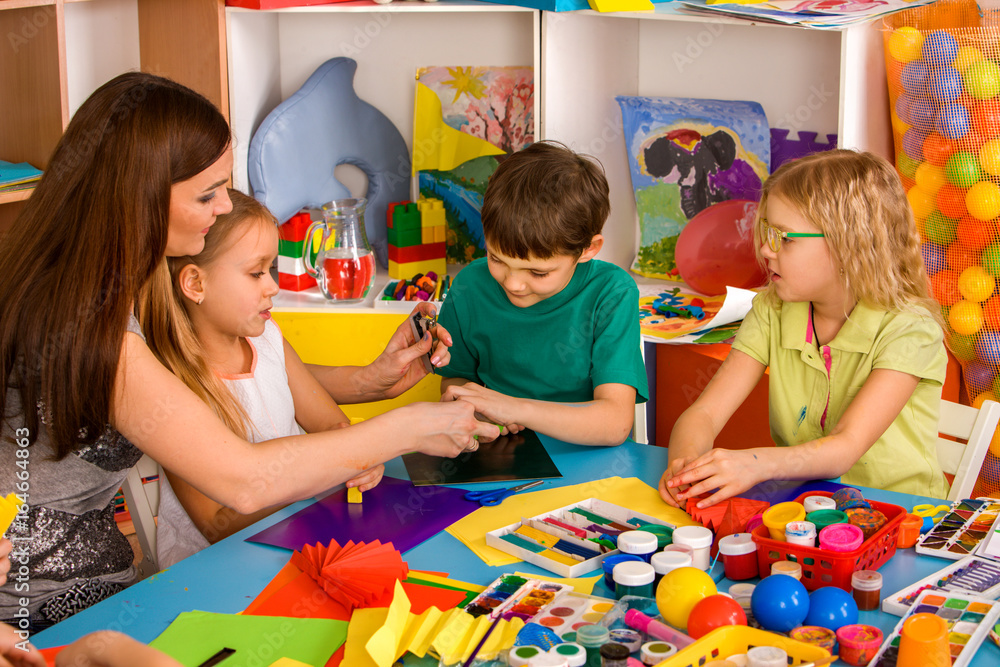 Naklejka premium School children with scissors in kids hands cutting paper with teacher in class room. Group of children in an orphanage. Large group girls and boys together.