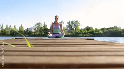 Young woman sitting on a wooden bridge on a river in the Lotus position with her eyes closed. Yoga in nature. photo
