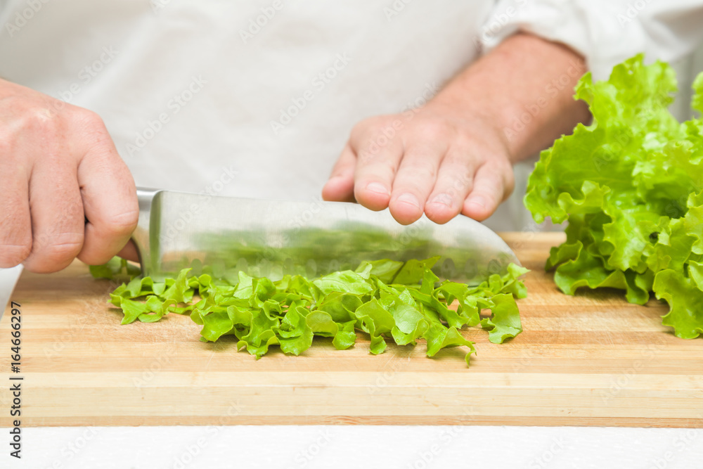 Chef's hands with knife cutting a green salad leaves on the wooden board. Preparation for cooking. Healthy eating and lifestyle.