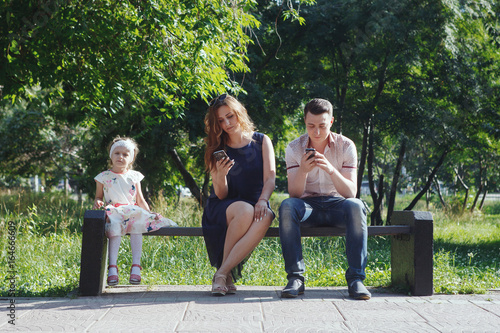 Young parents ignoring each other and the child, using their smartphones. In the summer the Park sitting on the bench © olgasparrow