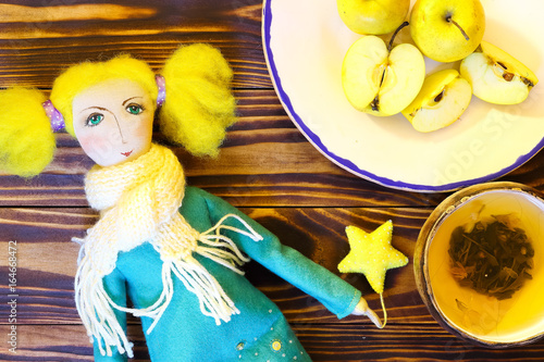 Soft handmade doll from textile of cute girl kolyada with bright yellow hair in coat and a scarf with asterisk in hand and cup of green tea and plate with sliced green apples on brown wooden boards. photo