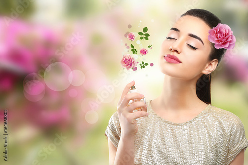 Beautiful young woman spraying perfume with flower component onto body on blurred background