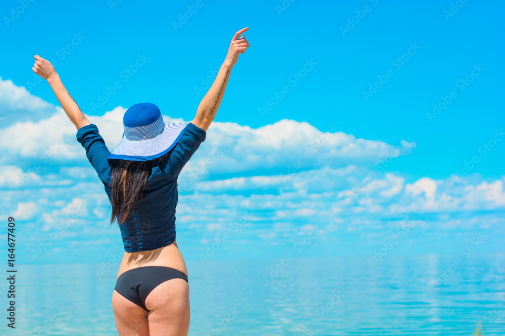 Obraz premium Gorgeous stylish brunette sexy woman on a beach in hat, mirror sunglasses and blue shirt rest near ocean and blue lagoon have a good mood in pretty day