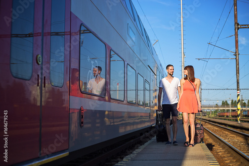 Happy young caucasian couple walking next to the train, pulling suitcases, wearing casual summer clothes