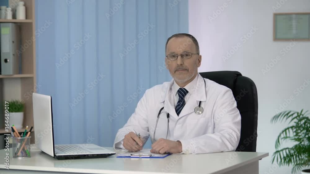 Doctor writing in patients medical record, looking into camera, clinic services Doctor writing in patients medical record, looking into camera, clinic services