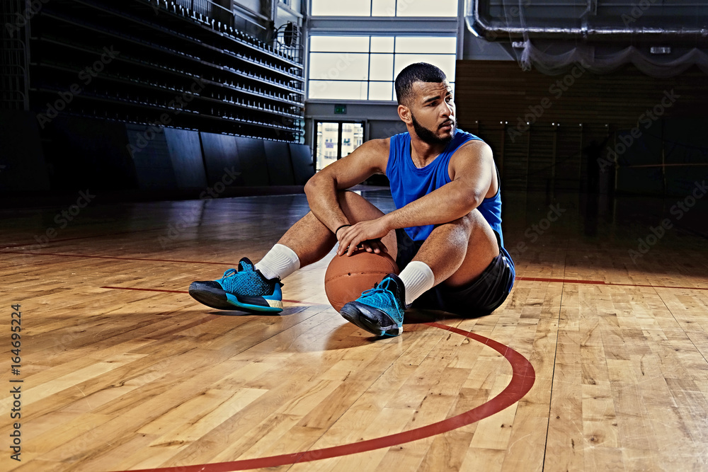 Fototapeta premium Black professional basketball player sits on a floor in a game hall.