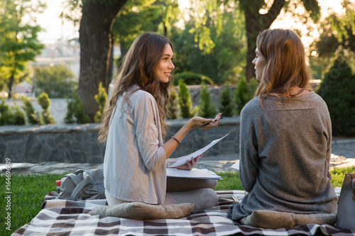 Smiling young two women sitting outdoors in park writing notes
