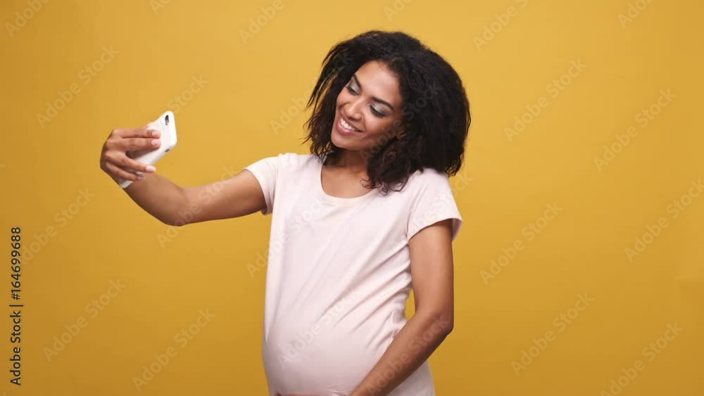 Happy afro american pregnant woman waving her hand while having video call conversation isolated over yellow background Happy afro american pregnant woman waving her hand while having video call conversation isolated over yellow background