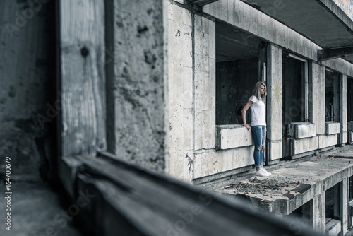 A young girl in a white T-shirt and blue jeans is standing on the edge of a ruined balcony in a ruined building early in the morning. © Artem Markin