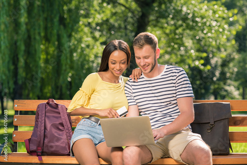 Young multiethnic couple shopping online with laptop and credit card while sitting on bench in park © LIGHTFIELD STUDIOS