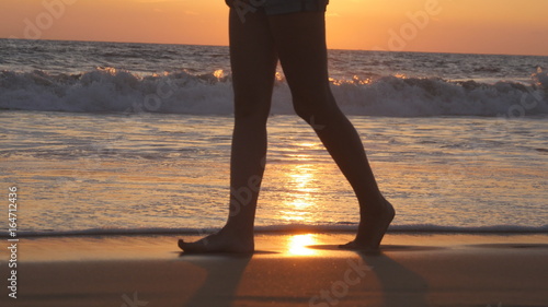 Legs of young woman going along ocean beach during sunrise. Female feet walking barefoot on sea shore at sunset. Girl stepping in shallow water at shoreline. Summer vacation concept. Close up