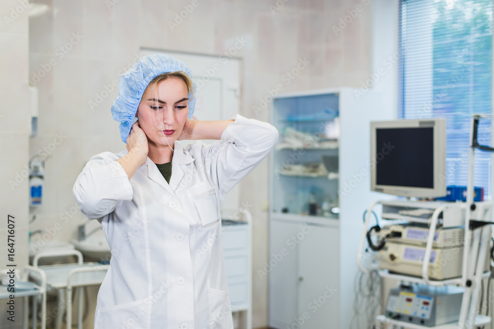Young doctor during preparation for operation. Assistant of surgeon wearing medical cap in operation room at the hospital. Female surgeon attaching his mask in an operating theater. Young doctor during preparation for operation. Assistant of surgeon wearing medical cap in operation room at the hospital. Female surgeon attaching his mask in an operating theater.