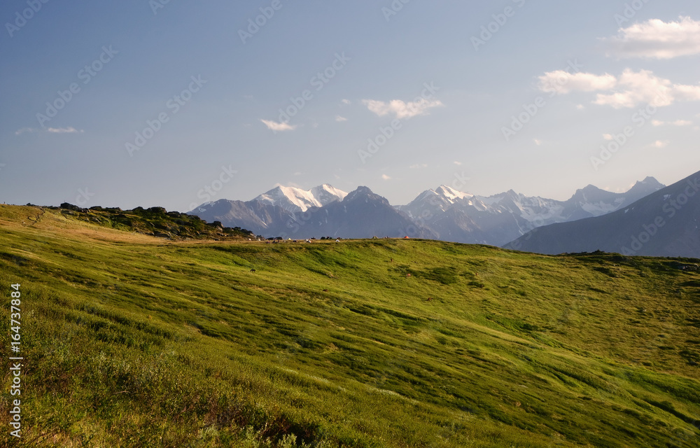 Fototapeta premium Mountain ranges and snow peaks with alpine meadow hiking camp of tents in the foreground under blue sky and white clouds Altai Mountains Siberia, Russia