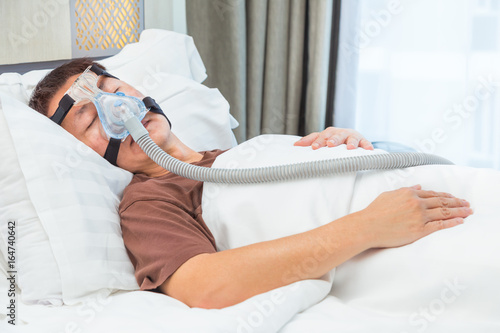 middle age asian man sleeping in his bed wearing CPAP mask connecting to air hose, device for people with sleep apnea photo