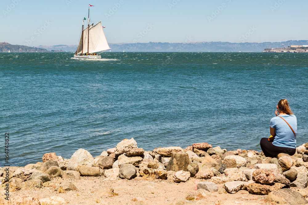 Woman sitting on beach gazing out at sailboat on water. Rocks in foreground, copy space in sky and water.