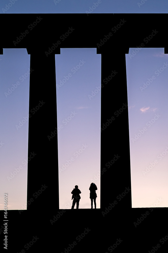 Fototapeta premium Silhouette of tourists standing amongst huge pillars of a monument taking photos of the sunset. Scottish National Monument, Edinburgh, Scotland, UK