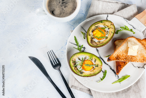 Healthy vegan breakfast. Diet. Baked avocado with egg and fresh salad from arugula, toast and butter. On a white marble plate, a light concrete table. A cup of coffee. Copy space top view