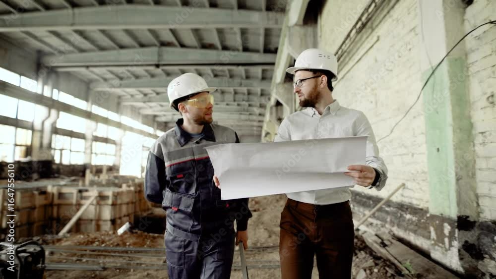 Auditor dressed in shirt and trousers walking with worker along building area. Professional inspector is holding blueprint and discussing detailes of future project with construction superintendent. Auditor dressed in shirt and trousers walking with worker along building area. Professional inspector is holding blueprint and discussing detailes of future project with construction superintendent.