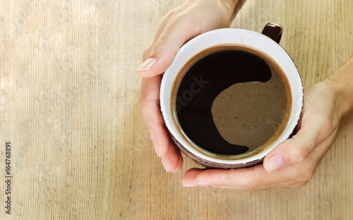 Hands of a woman hugging a large cup of coffee. Wooden background. Top view. Copy space
