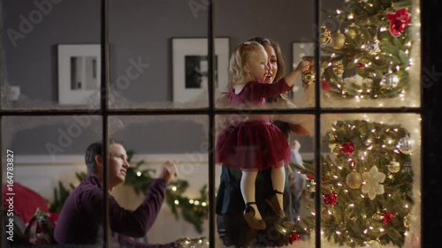 Medium zoom in shot of family decorating Christmas tree behind window / Cedar Hills, Utah, United States