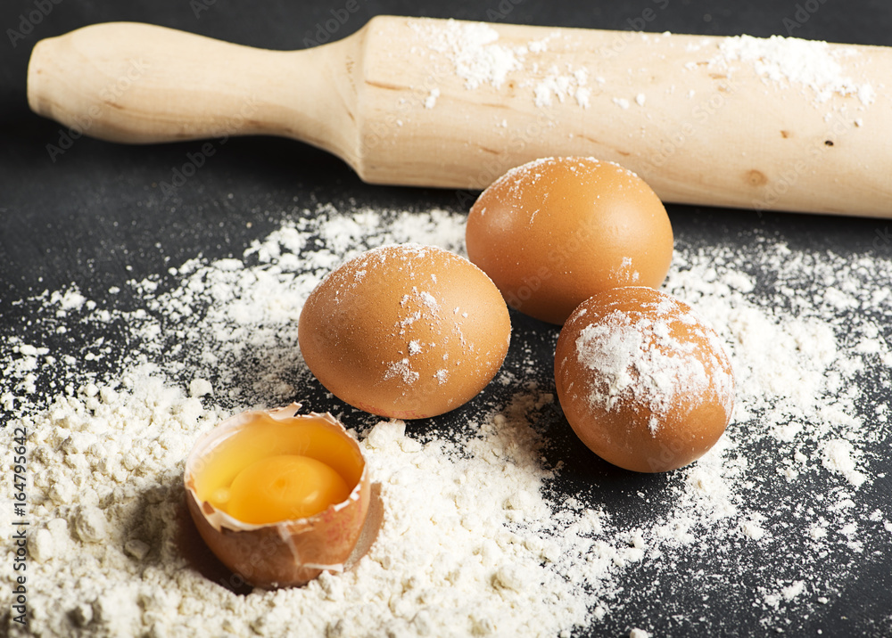Eggs on flour next to wooden roller on black wooden table. Horizontal studio shot. Eggs on flour next to wooden roller on black wooden table. Horizontal studio shot.