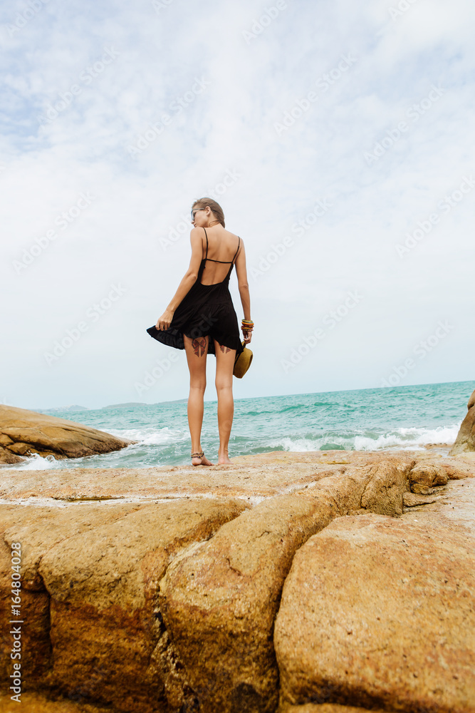 Sexy fit lady on big rocks correct her little black summer dress flying in the wind. View from back. Beauty cute girl on a tropical beach sea ocean shore with large stones. Outdoor summer lifestyle.