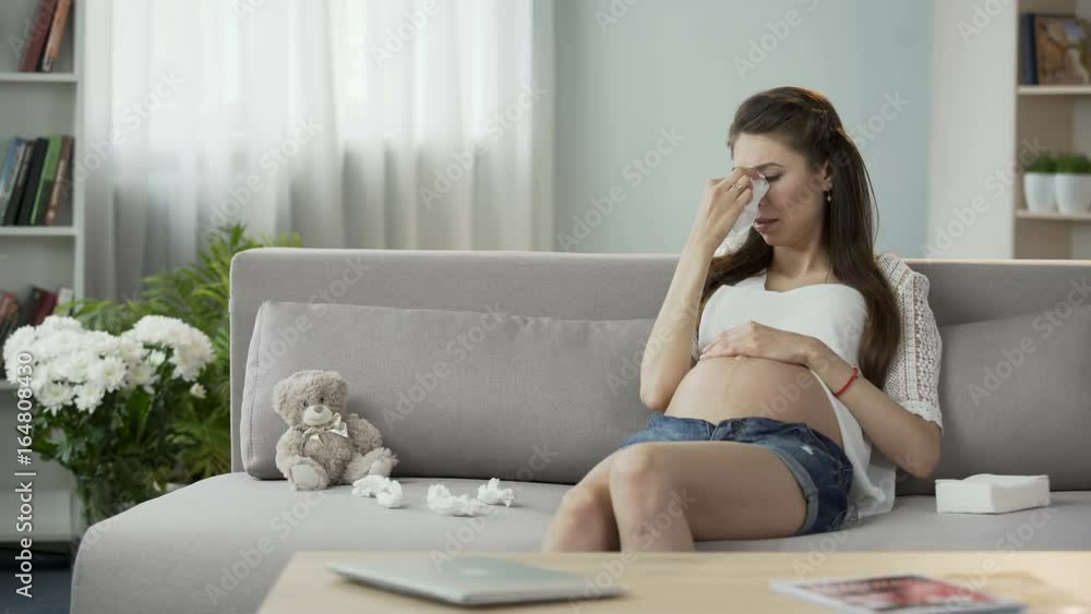 Pregnant lady sitting on sofa crying drying tears with tissue, hormonal disorder Pregnant lady sitting on sofa crying drying tears with tissue, hormonal disorder