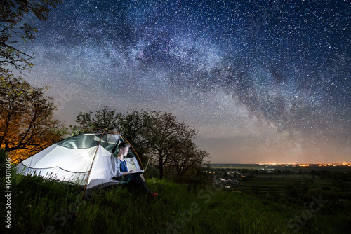 Woman using her laptop in the camping at night. Woman sitting in the tent under beautiful night sky full of stars and milky way. On the background luminous village. Long exprose