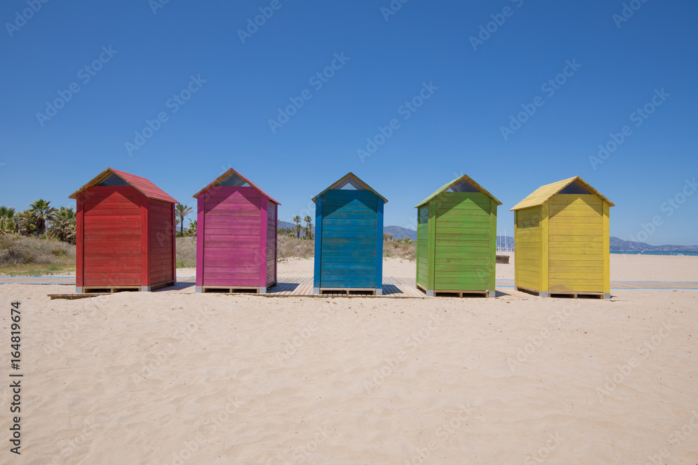 five wooden bathing cabins or huts, colored in blue, red, pink, yellow and green, and wooden footway on sand, Beach of PIne or Pinar, Grao Castellon, Valencia, Spain, Europe. Blue sky
