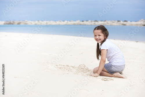 Happy child on the beach. Paradise holiday concept, girl seating on sandy beach with blue shallow water and clean sky