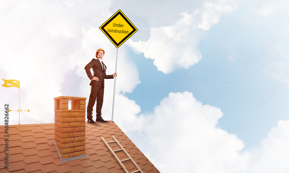 Young businessman on house brick roof holding yellow signboard. Mixed media Young businessman on house brick roof holding yellow signboard. Mixed media