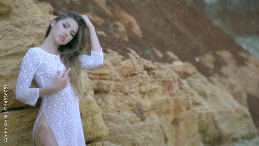Beautiful blonde with long hair in a white translucent dress stroking herself standing among rocks. Slow motion shooting Beautiful blonde with long hair in a white translucent dress stroking herself standing among rocks. Slow motion shooting