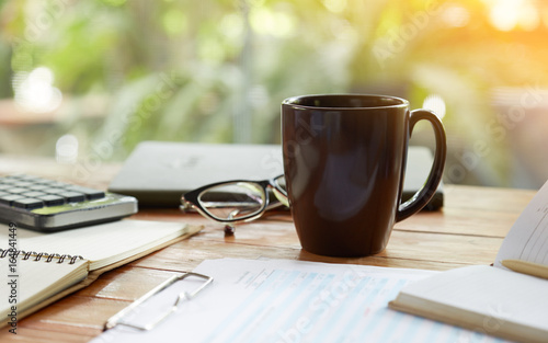 Coffee mug and business objects on wooden desk with greenery background, selective focus