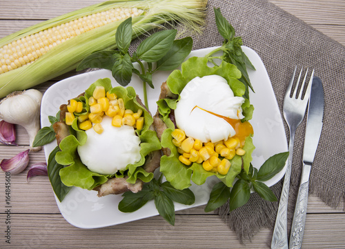 Poached eggs in the basket of bacon with herbs and tomatoes and corn with garlic. On wooden background.