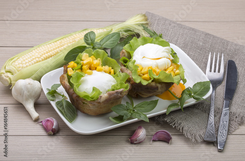 Poached eggs in the basket of bacon with herbs and tomatoes and corn with garlic. On wooden background.