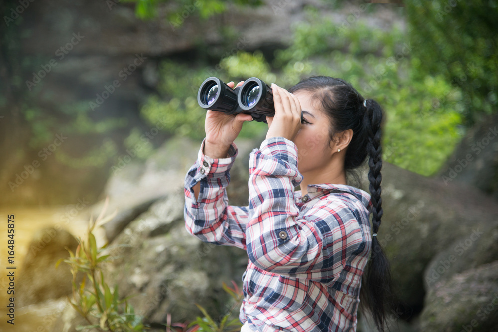 Young Asian traveler woman looking a binoculars in happiness and sitting on a rock of waterfall.