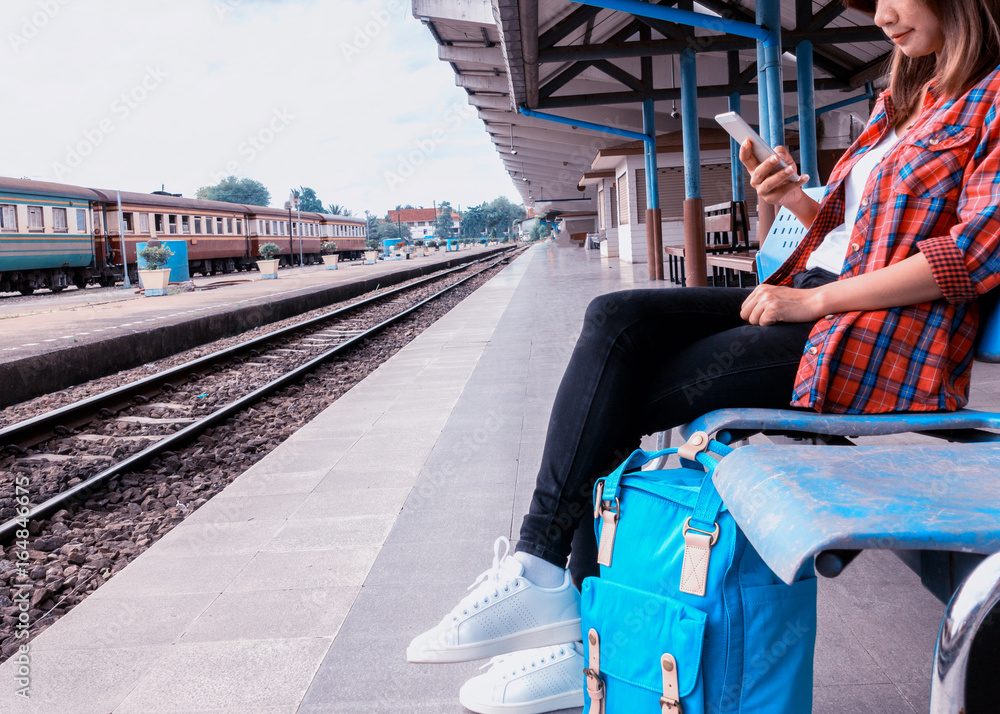Young woman traveler and backpack waiting railway at train station, Young woman sitting with using smartphone at the train station, Summer holiday and travel concept.