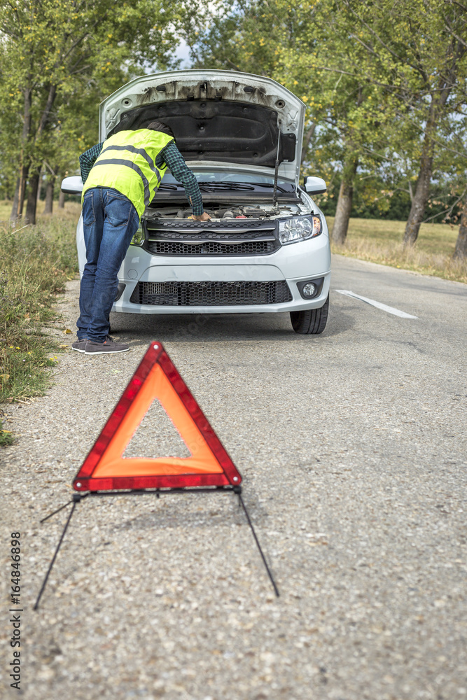 Young man examining his damaged car parked on the side of the road and a red triangle to warn