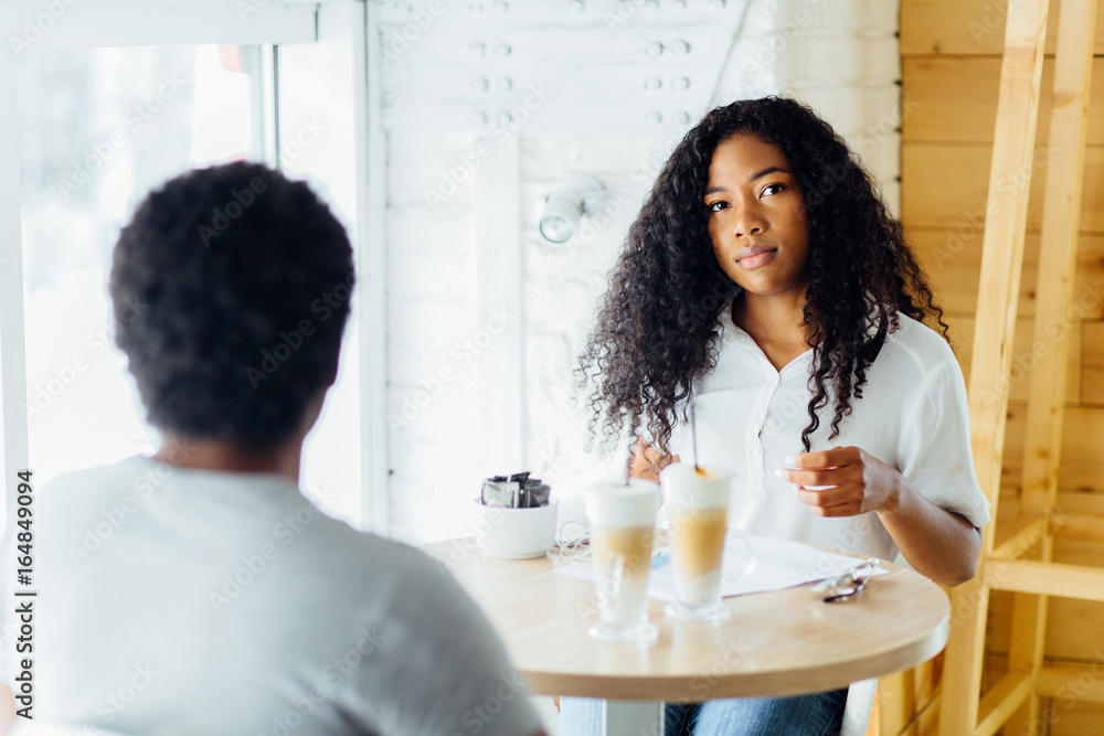 Black girl with Afro hairstyle holding a cup of coffee, listening music with headphones and looking at camera. Her boyfriend sitting with back to camera. Loving couple enjoying time together in cafe Black girl with Afro hairstyle holding a cup of coffee, listening music with headphones and looking at camera. Her boyfriend sitting with back to camera. Loving couple enjoying time together in cafe