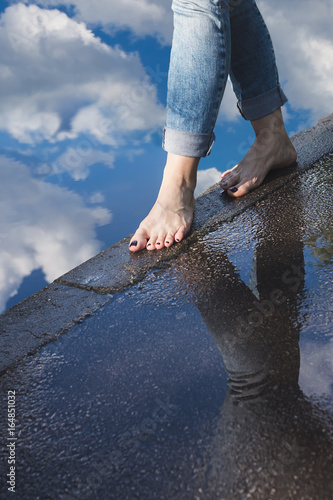 Barefoot woman in blue jeans walking on a water edge on a wet pavement