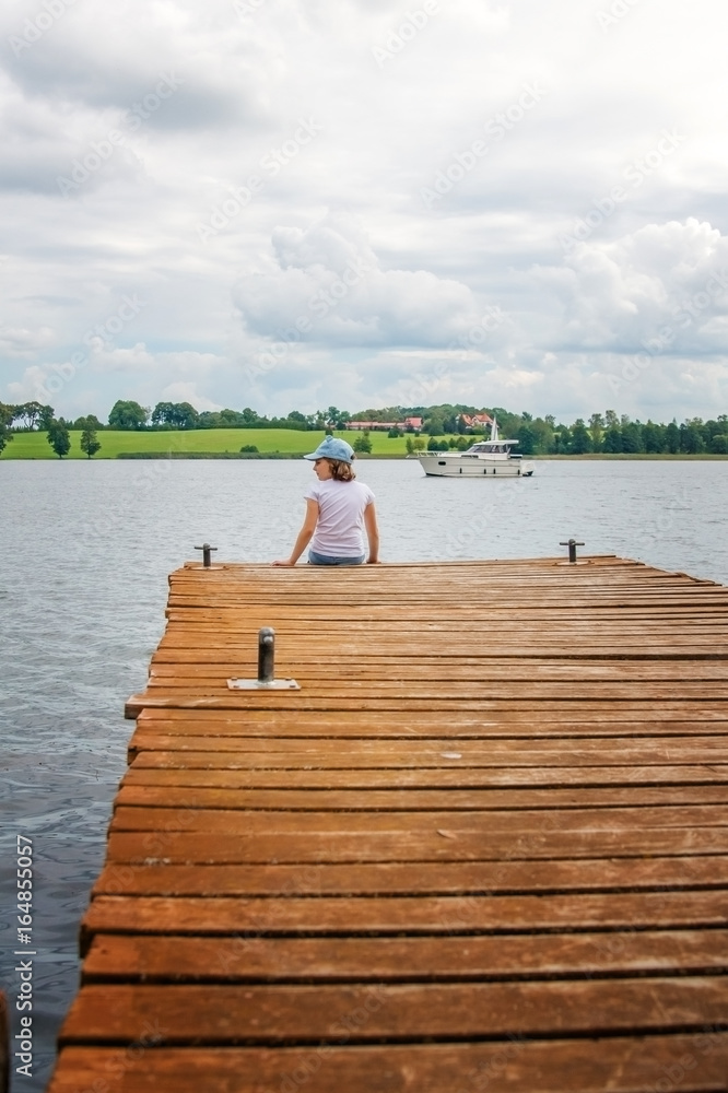 The child sits on a wooden pier on the river and looks at the water.